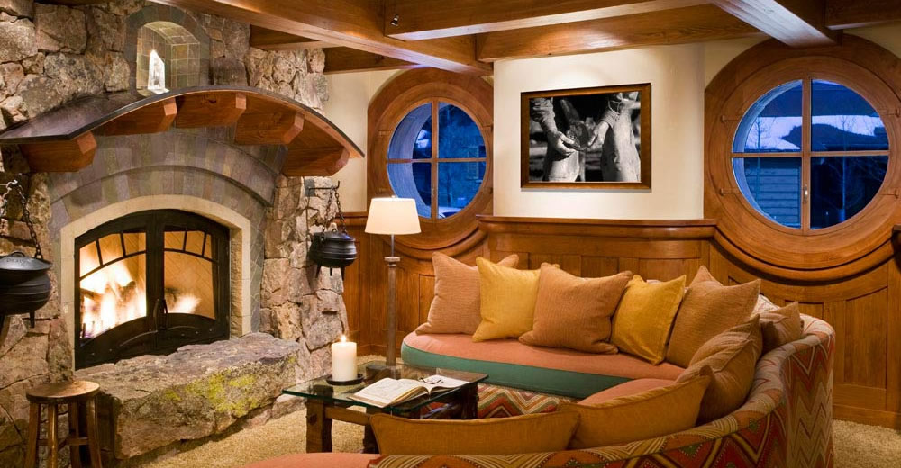 A living room with a stone fireplace with a curved mantel, wood paneling and round windows